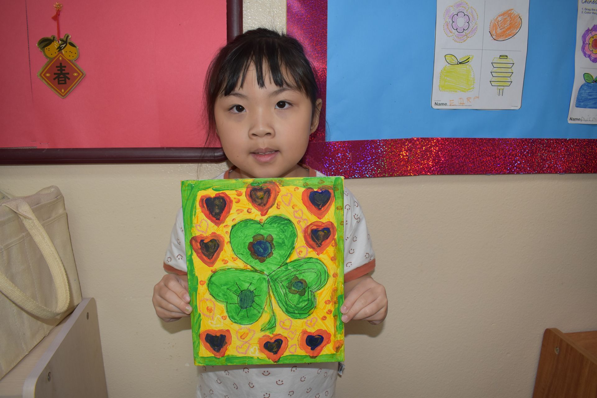 Girl holding artwork with a large green shamrock and red hearts on a yellow background.