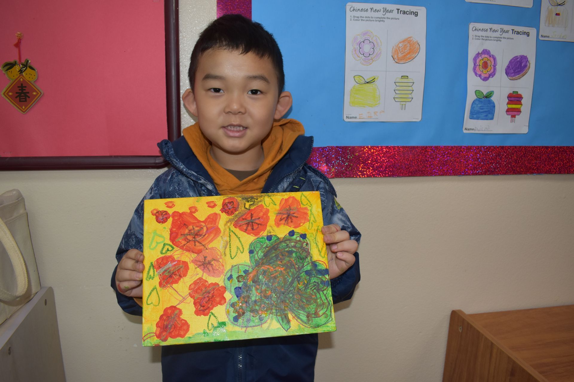 Young boy holding artwork, yellow and orange colors. Artwork on a bulletin board behind him.