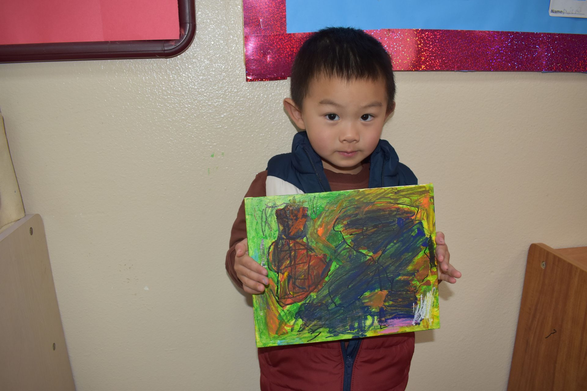 Young child holding a colorful abstract painting in a classroom setting.