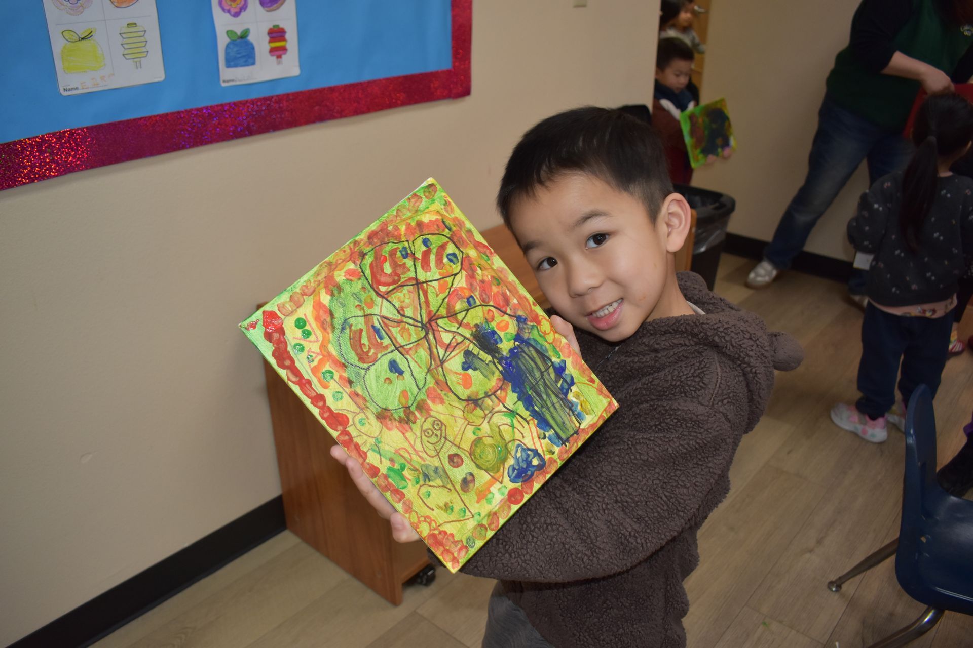 Young boy smiles, holding colorful artwork. Brown sweater, beige wall, classroom setting.