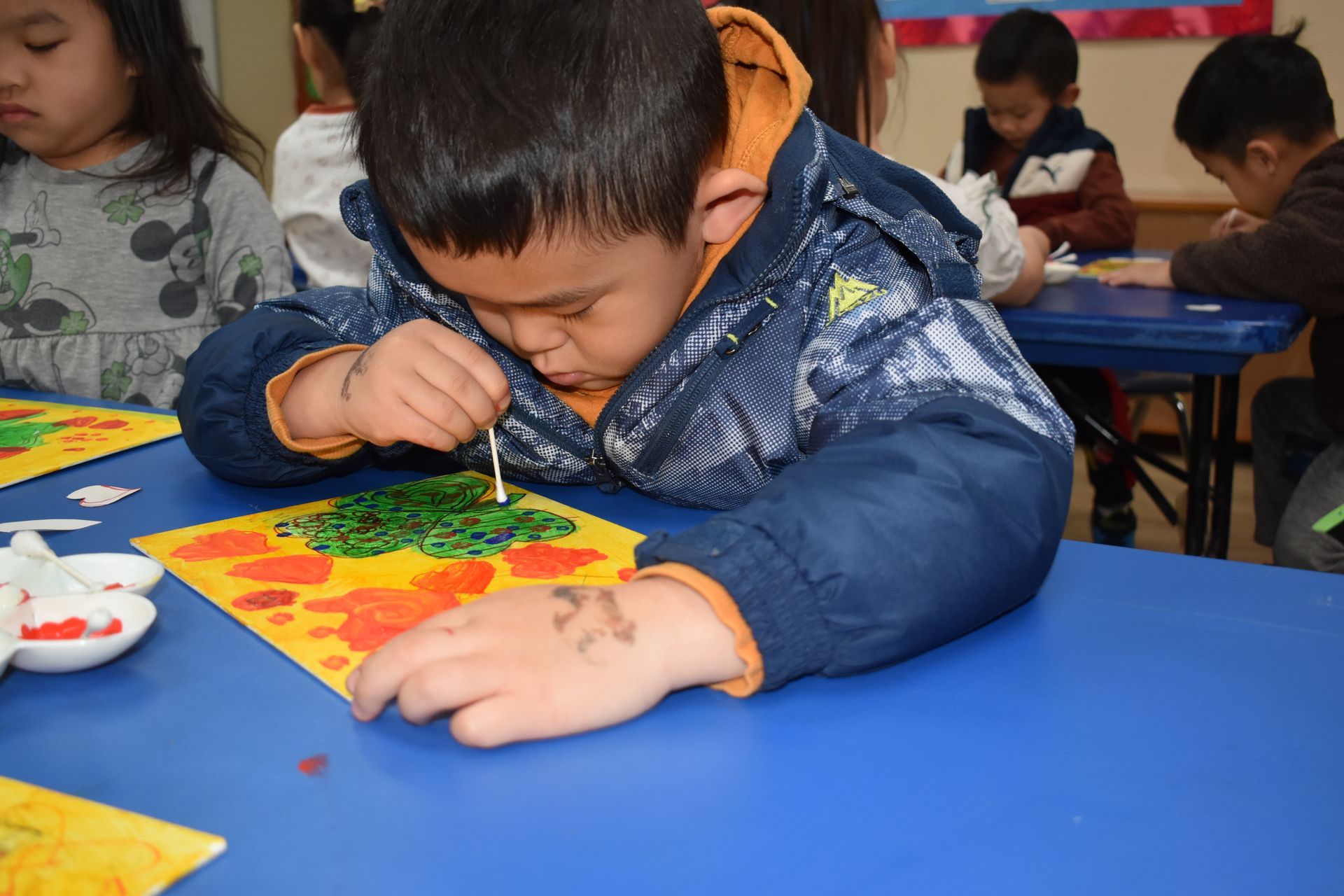 Boy in blue jacket using a paintbrush on artwork at a blue table in a classroom.