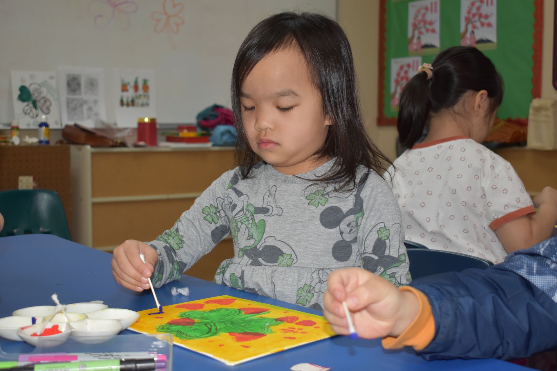 Child painting with a cotton swab at a table in a classroom.
