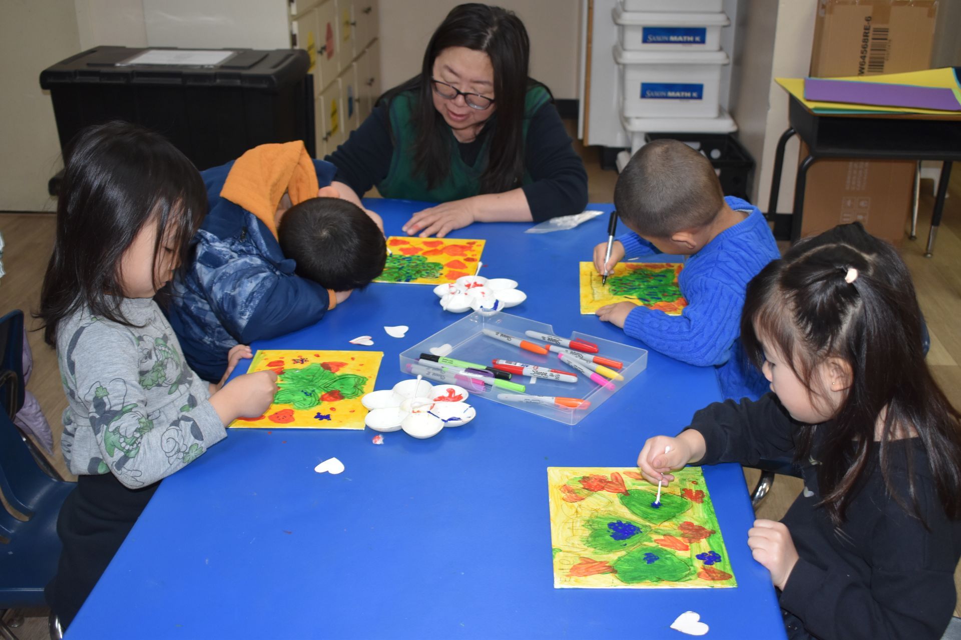 Children and an adult creating art at a table. They are using paint and markers on their projects.