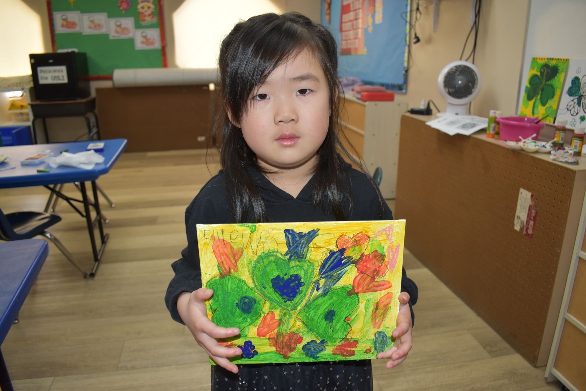 Young child holding up colorful artwork in a classroom setting.