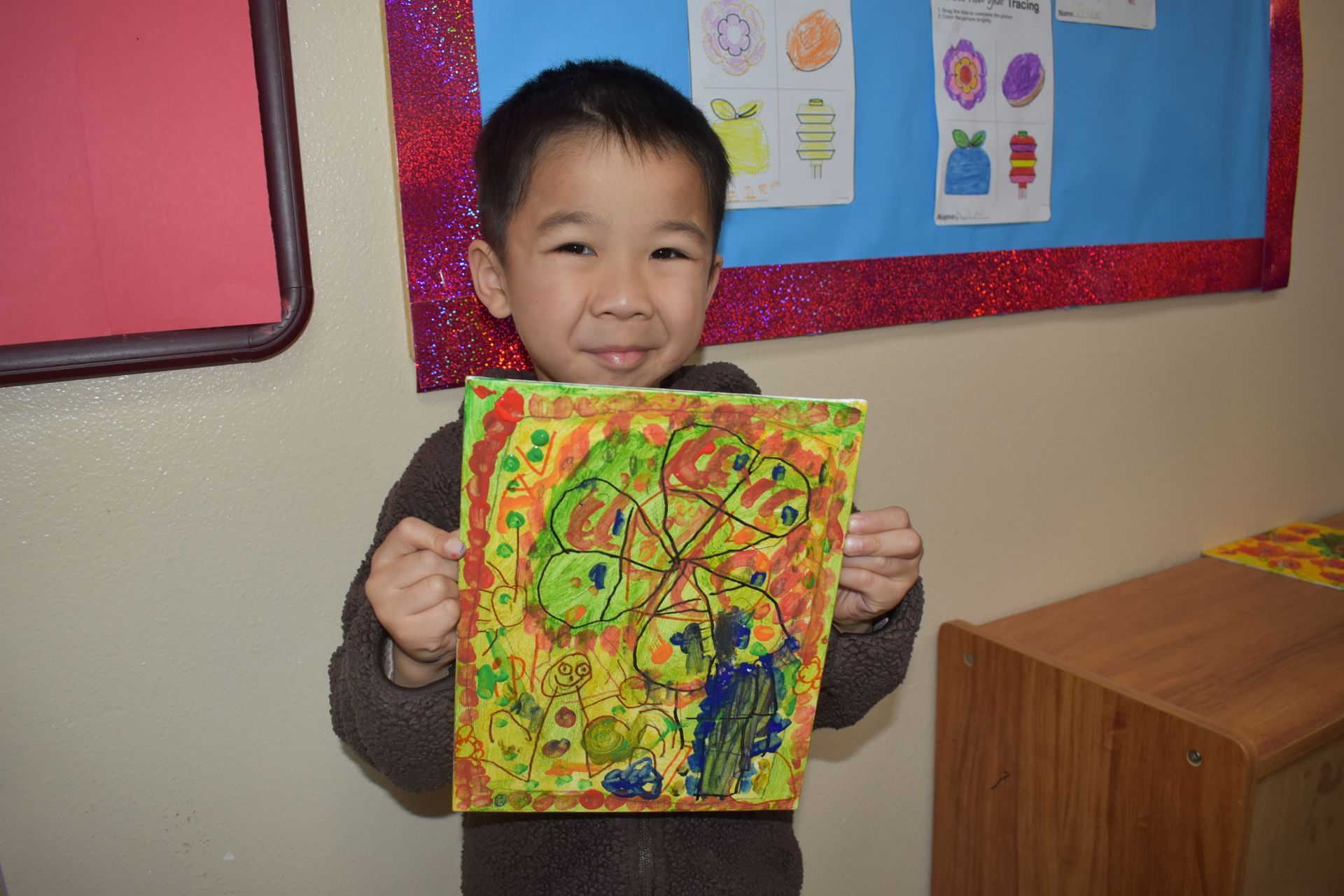 Boy smiles, holding a colorful abstract painting. He's inside, likely a classroom, with artwork on the wall.