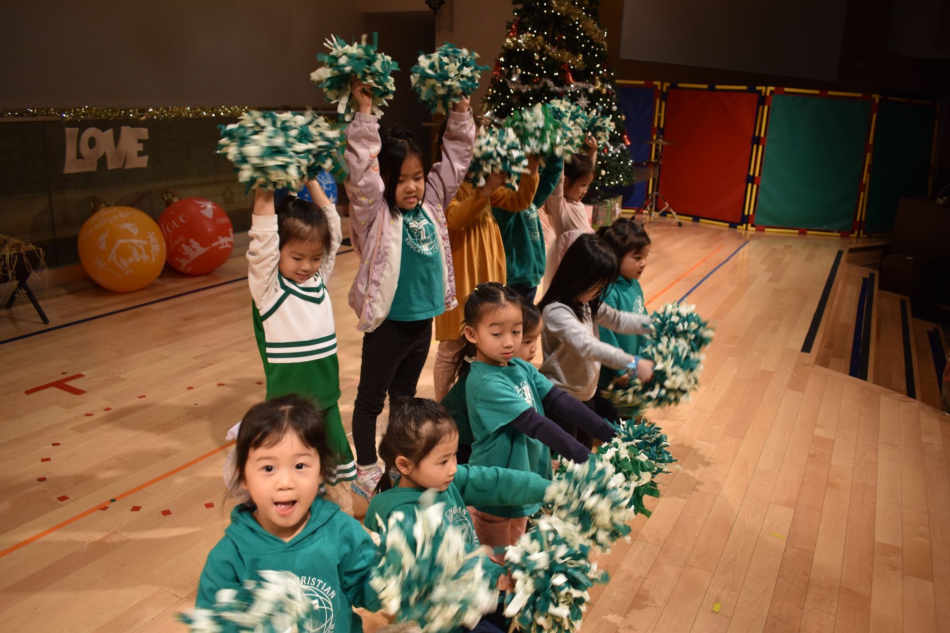 Children cheerleading with green pom-poms on a wooden floor, Christmas tree in background.