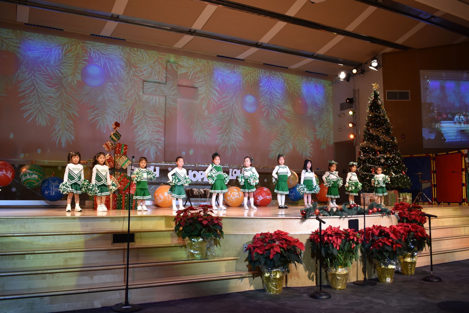 Children in green and white costumes performing on a stage decorated for Christmas, with a Christmas tree and projections.