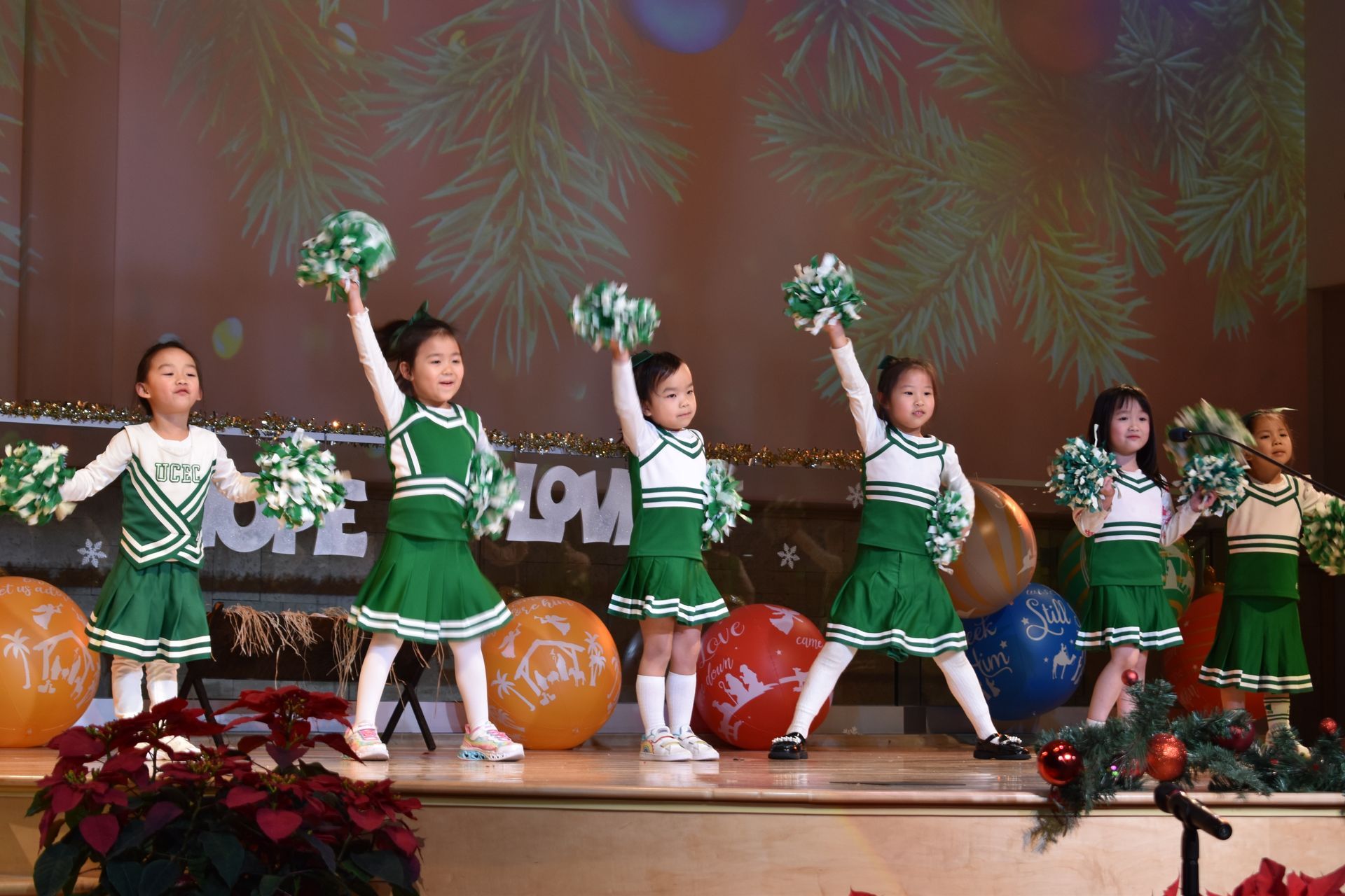 Five young cheerleaders in green and white uniforms perform on stage with pom-poms.
