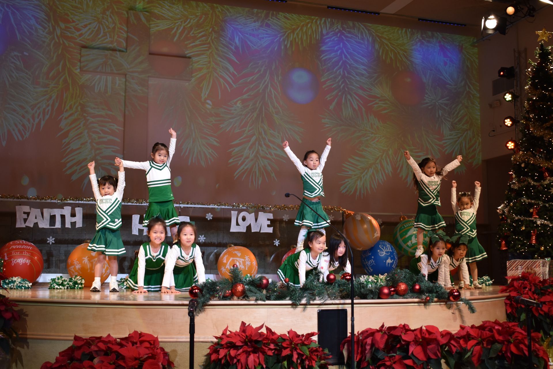 Children performing a cheerleading routine on a stage decorated for Christmas; green and white uniforms, arms raised.