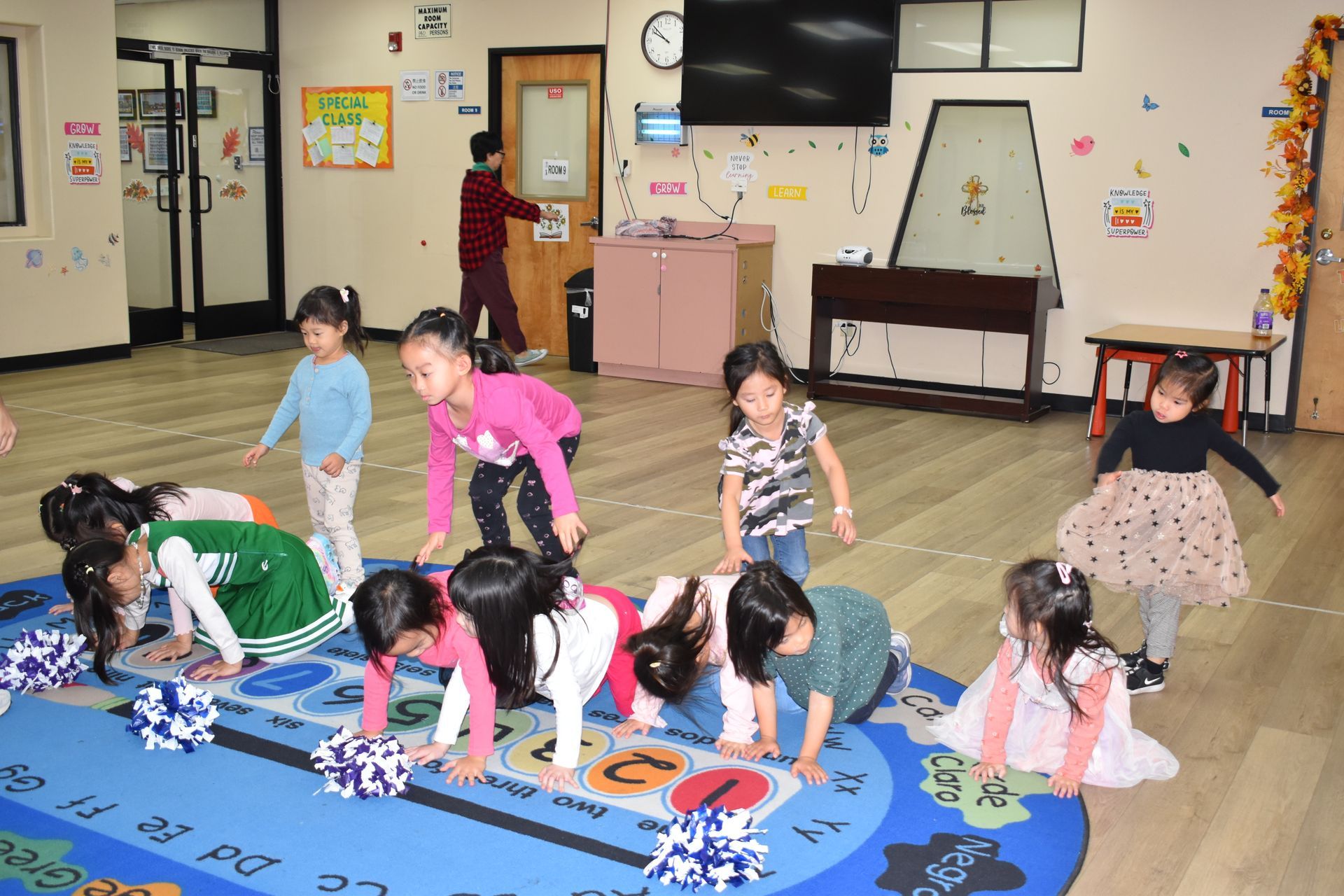 Children in a classroom, some with pom-poms, doing an activity on a colorful rug. A person opens a door in the background.