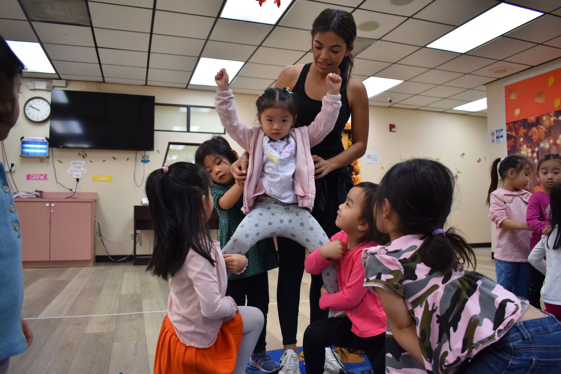 Teacher helps a child jump as other children watch in a brightly lit classroom.