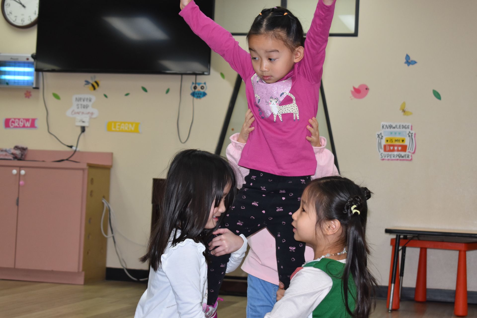 Three children playing: one held up with arms raised, others looking up. Indoors.