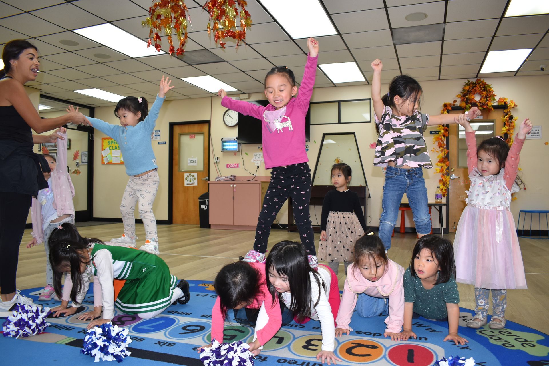 Children and instructor doing an exercise routine in a brightly lit room. Some children have arms up or bent down.