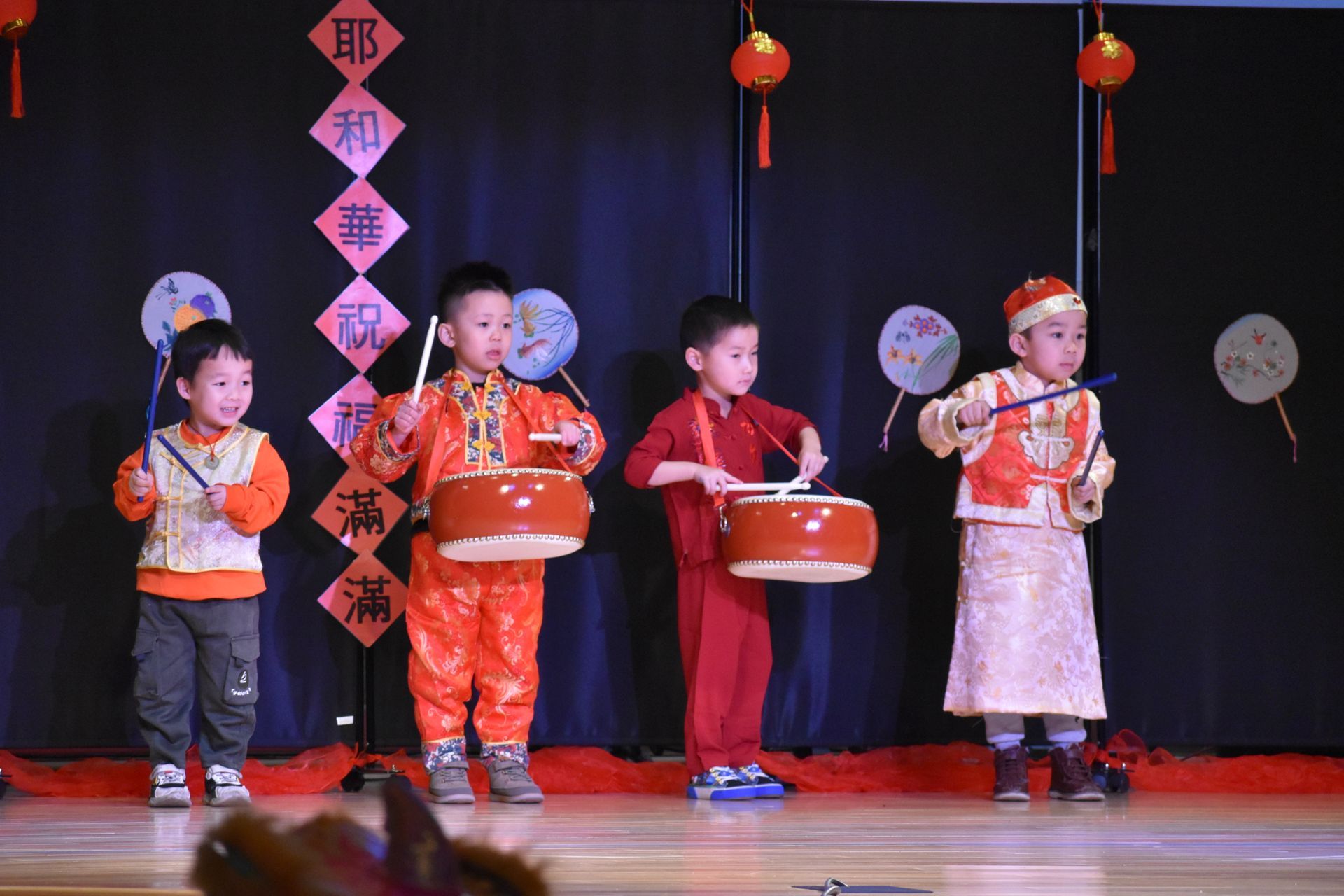 Four children in festive outfits playing drums on a stage with Chinese decorations.