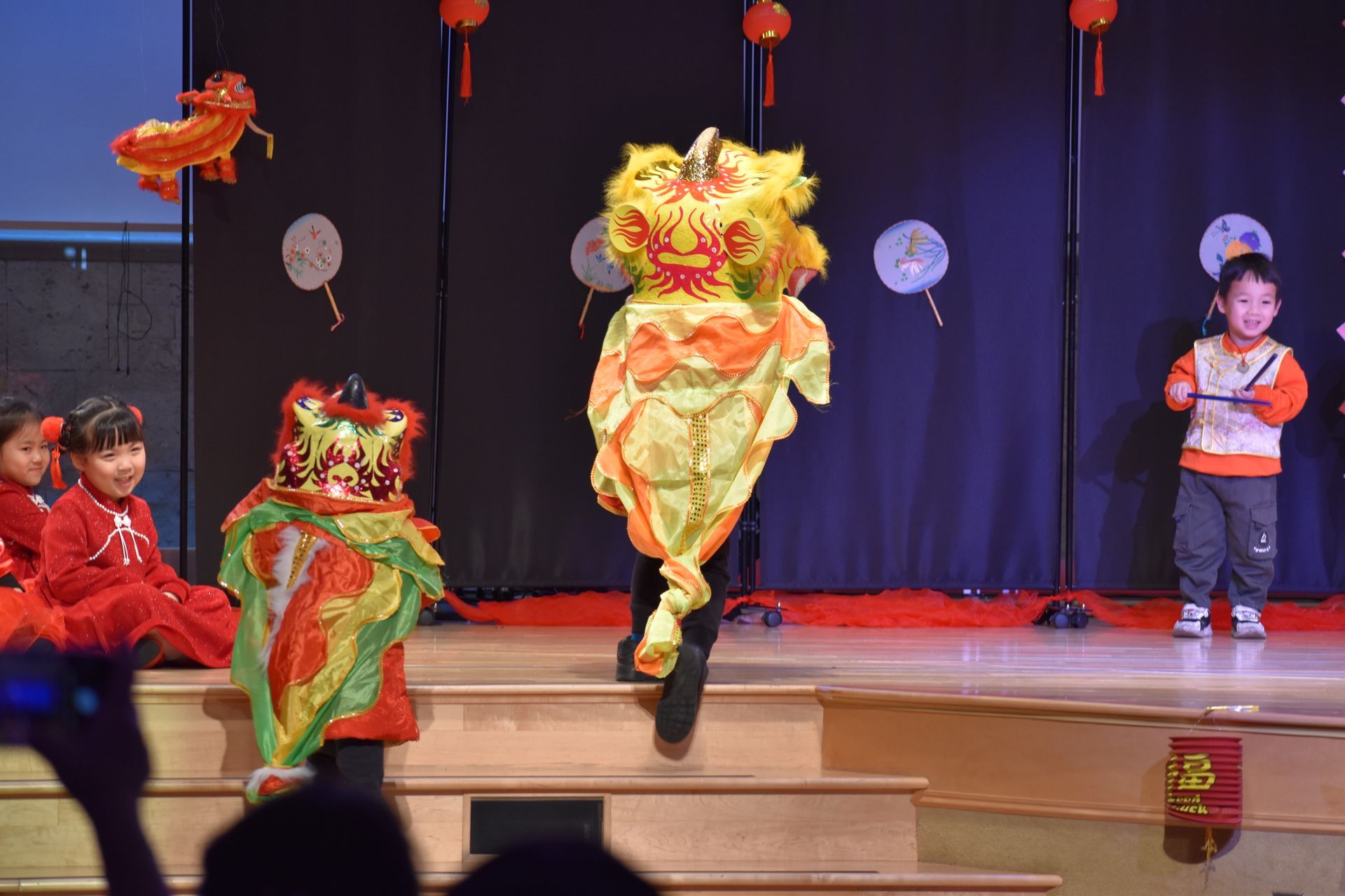 Children in lion dance costumes perform on a stage decorated for a festival.