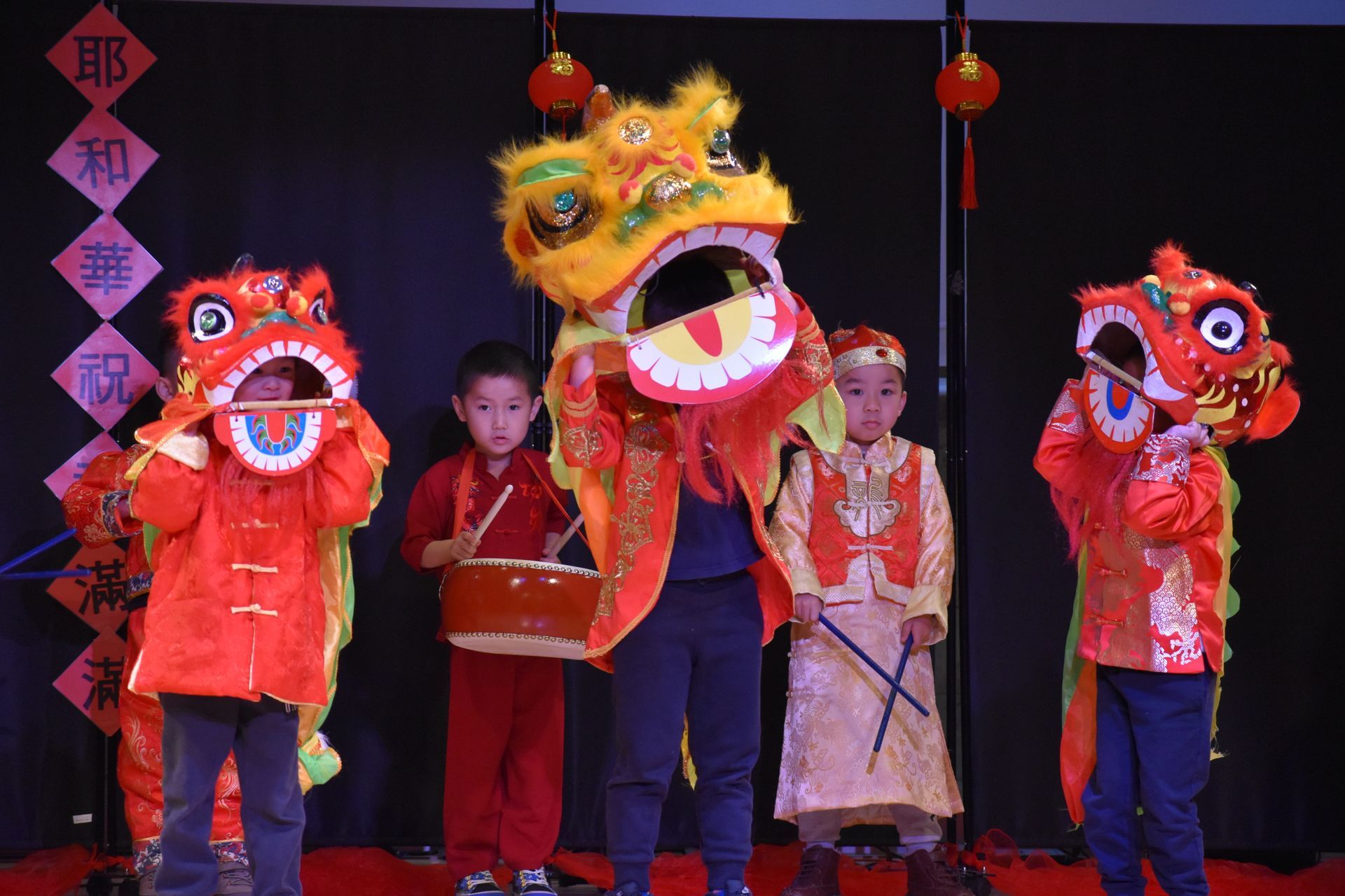 Children performing a Chinese lion dance on stage, wearing colorful costumes and holding instruments.