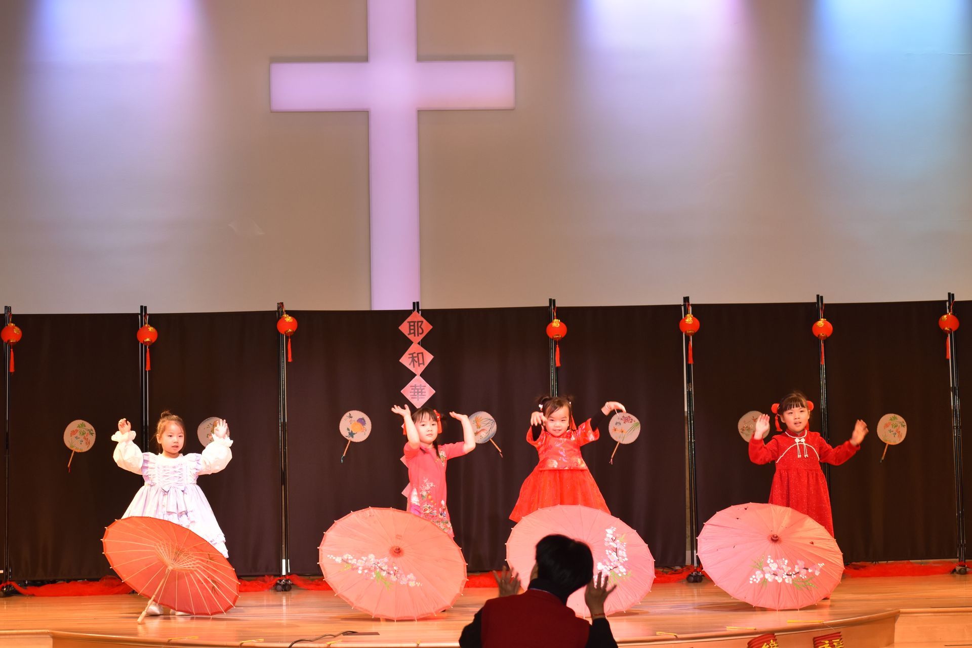 Children in red and white dresses dancing on stage with parasols and fans. Cross backdrop.
