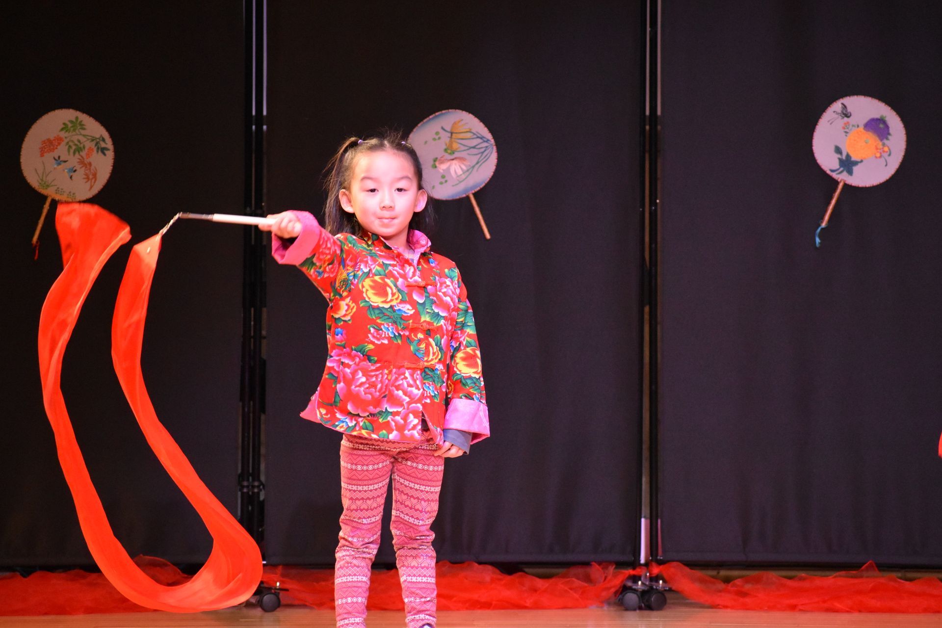 Child performer in red patterned outfit holding a red ribbon on a stage with decorations.