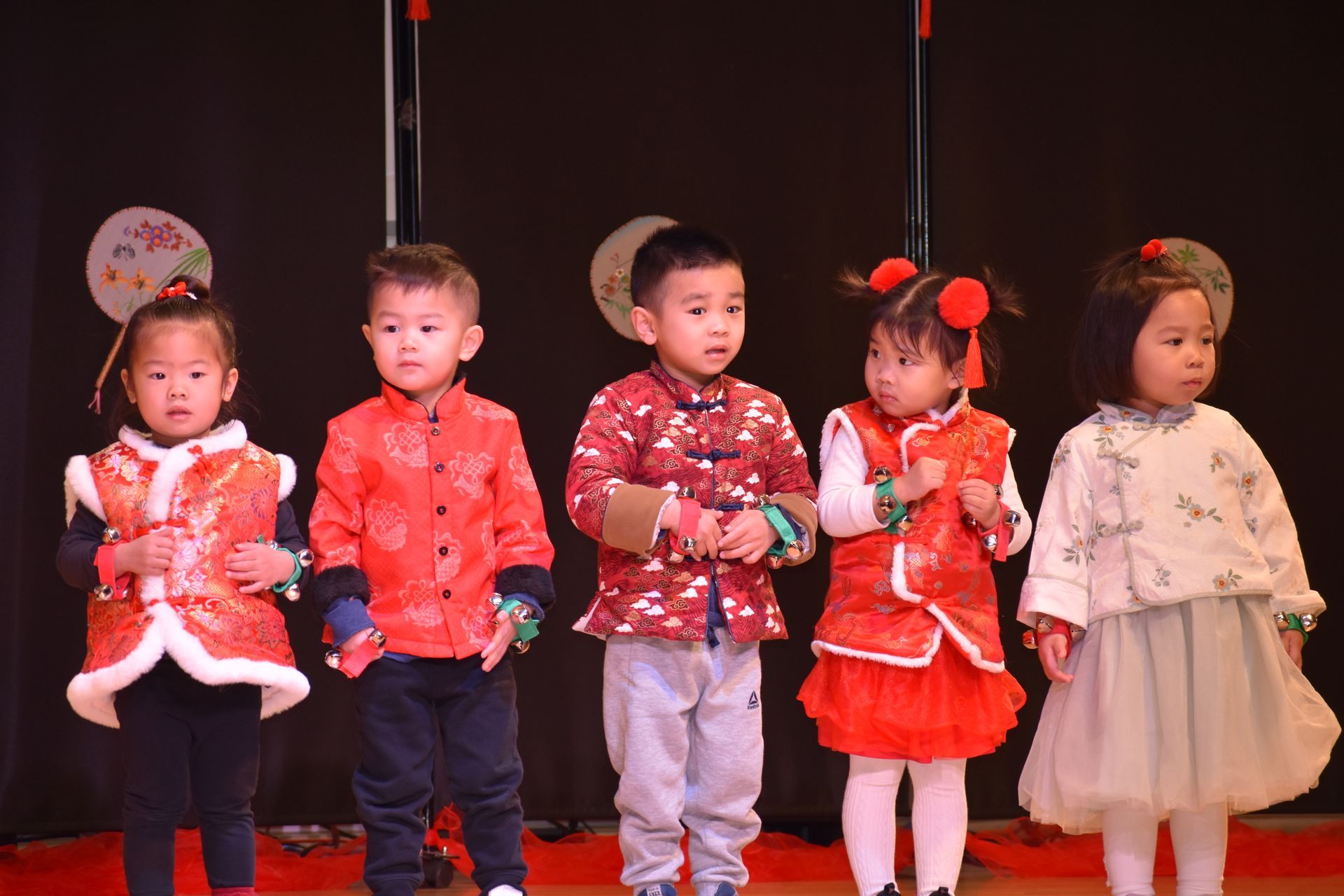 Five children in traditional attire stand on stage. Red and white garments. Stage with dark backdrop.