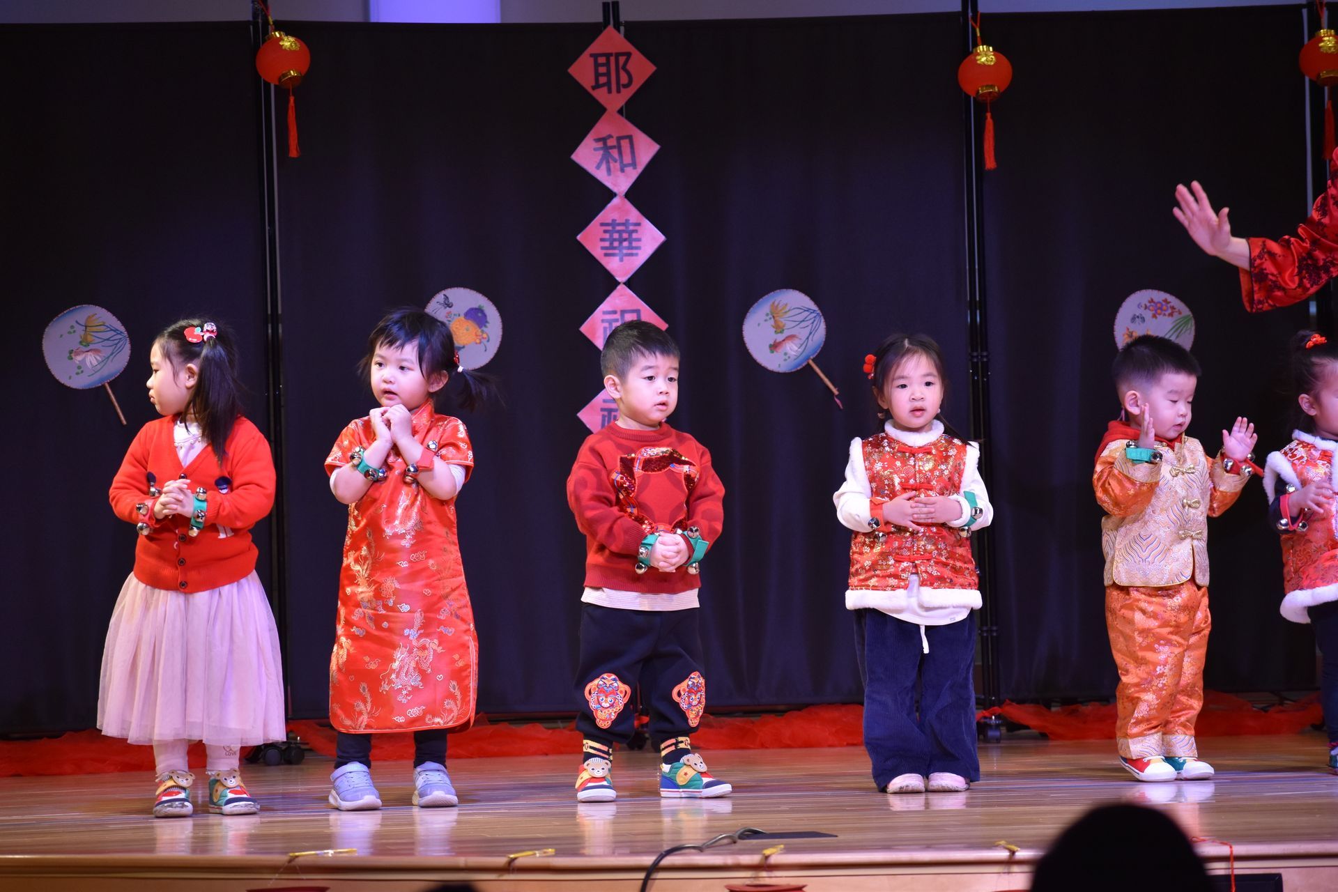 Children on stage in festive outfits perform. Stage has red lanterns and backdrop decor.