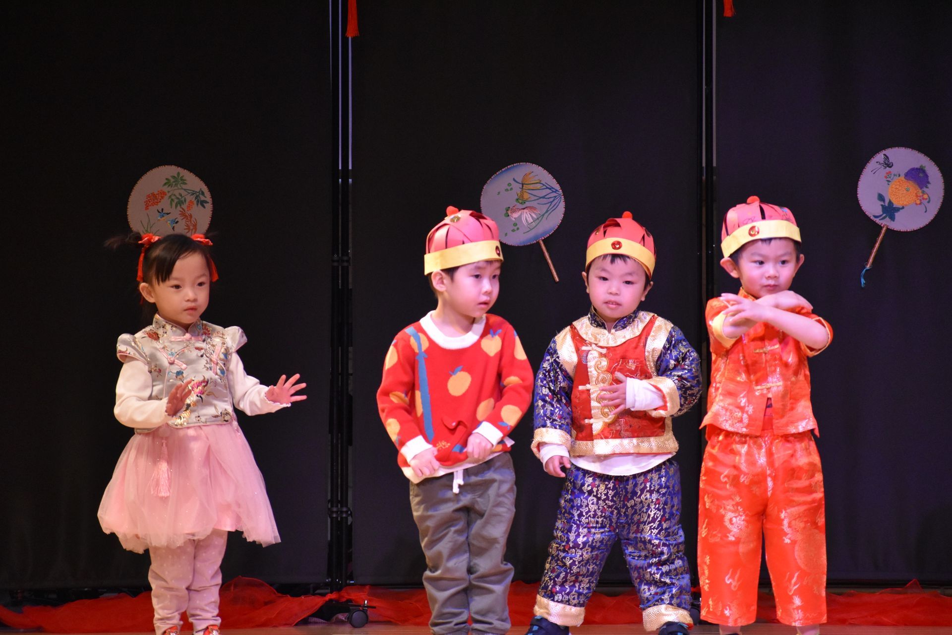 Children in festive outfits perform on stage; one in a pink dress, others in red/orange tops and hats, and blue pants.