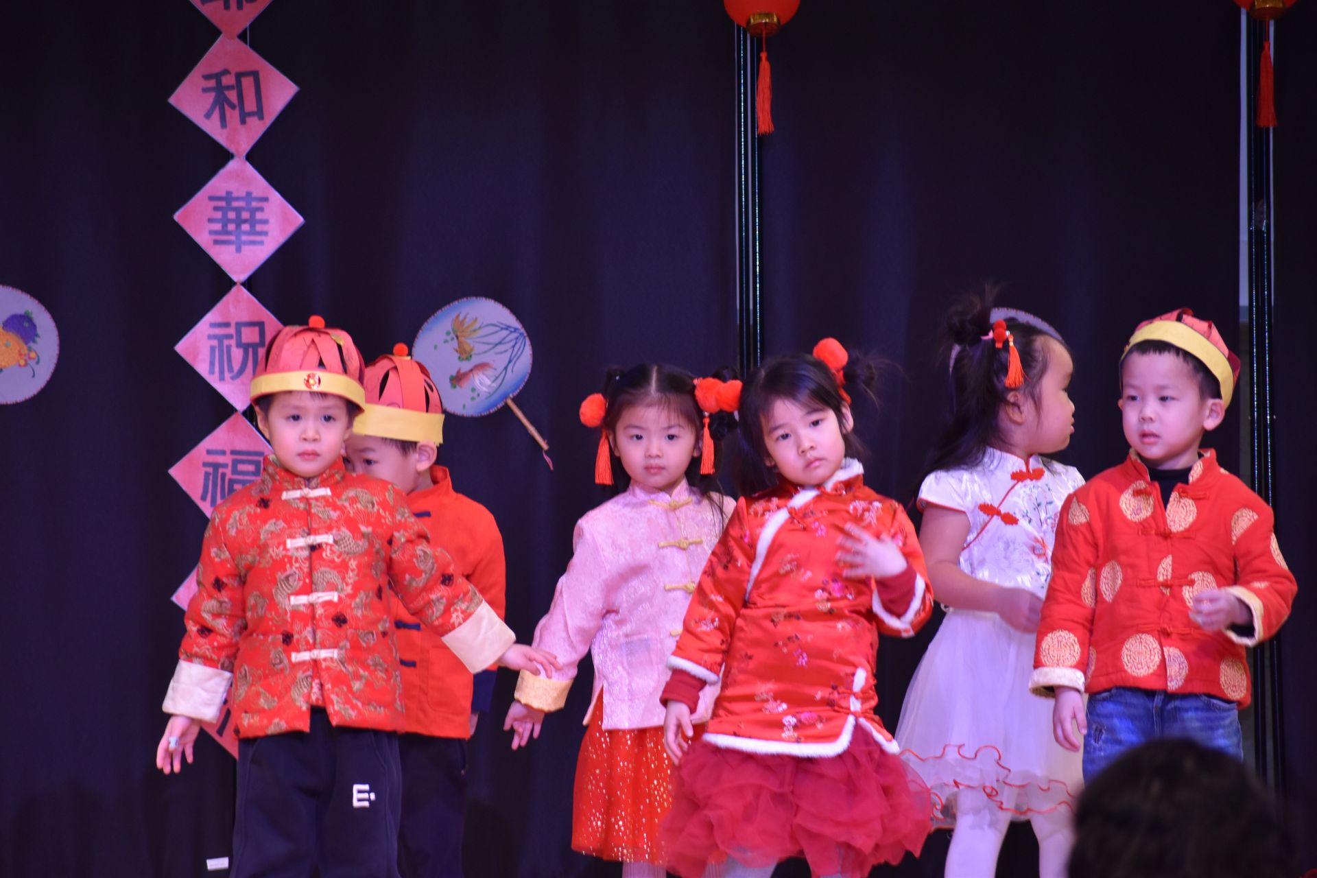 Children in red and pink traditional outfits on a stage with Chinese New Year decorations.