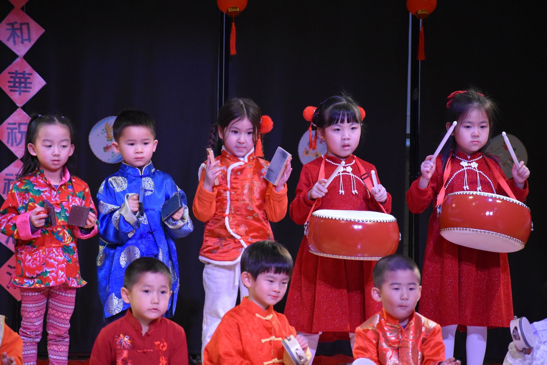 Children in traditional attire perform, playing drums and instruments on stage with lanterns.