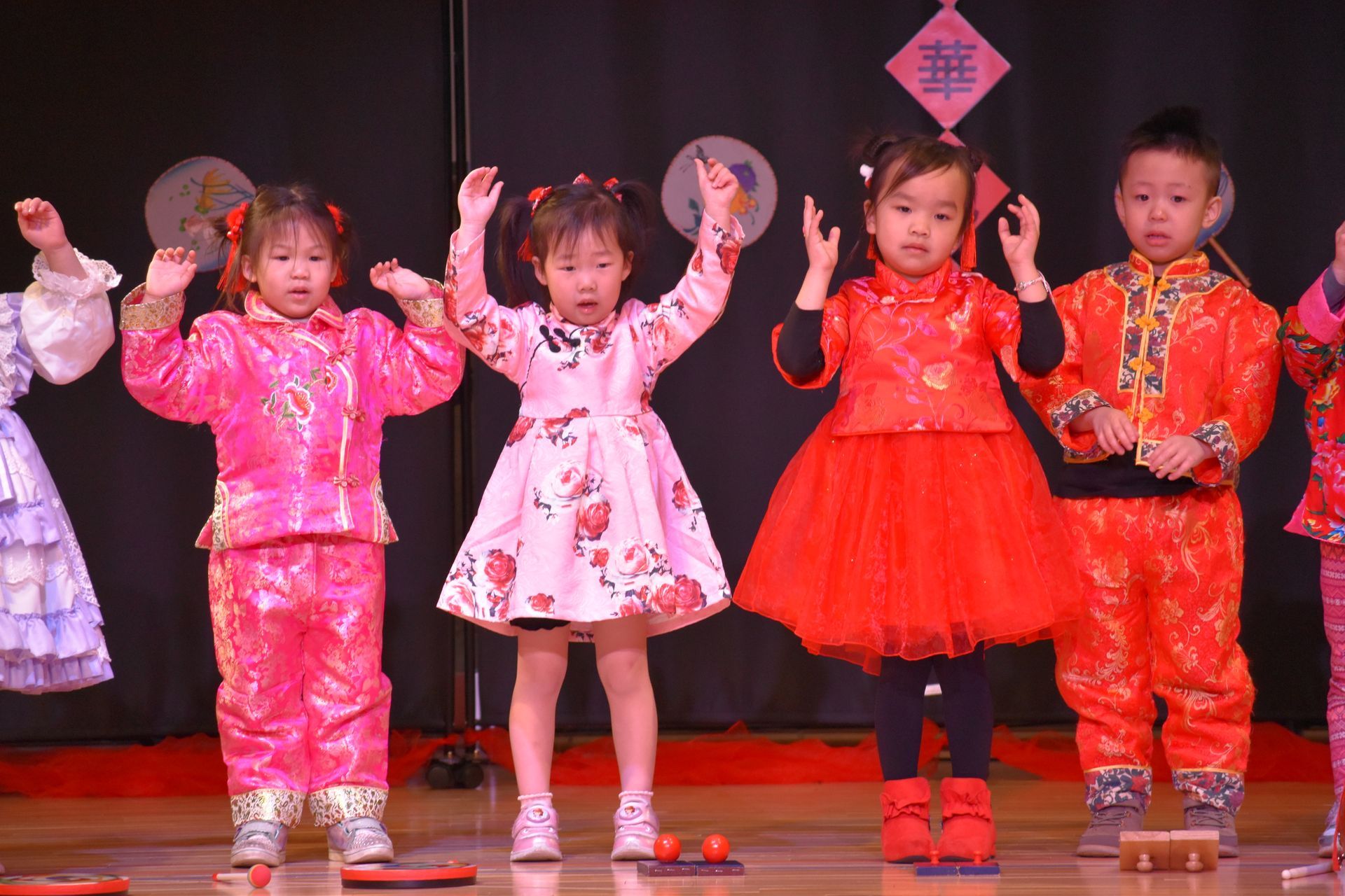 Children in colorful outfits on stage, arms raised. Red and pink clothing. Stage backdrop.