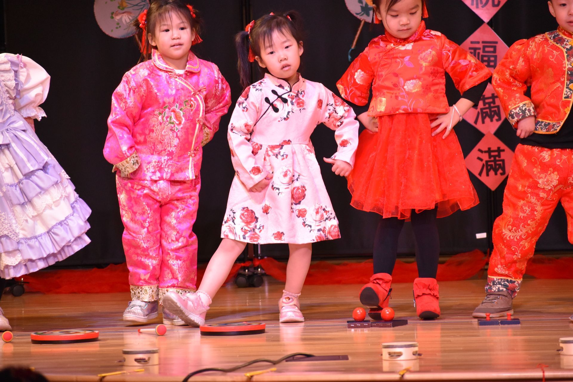 Children in festive outfits performing on stage, faces showing concentration, with props.