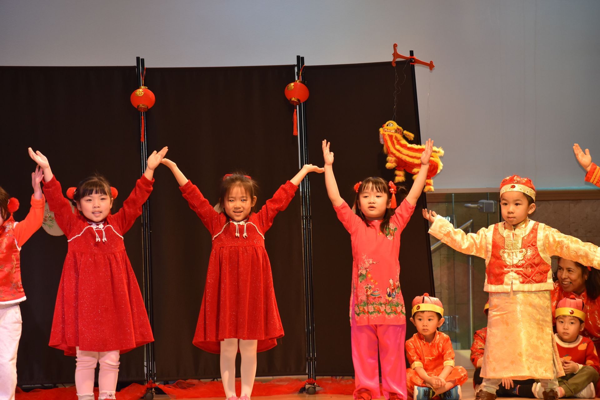 Children in red outfits performing on stage, arms raised. Stage has red lanterns and backdrop.
