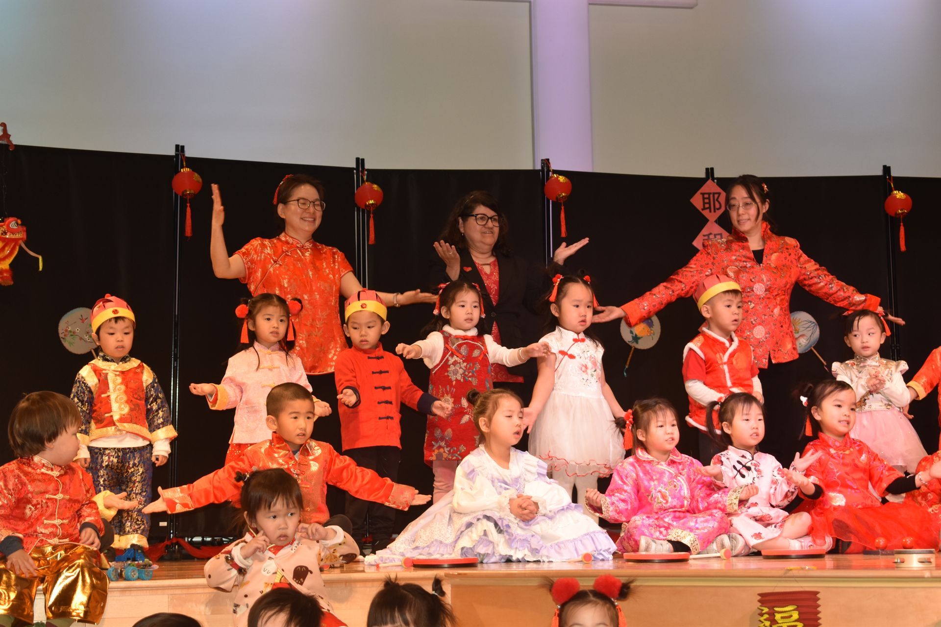 Children and adults in red outfits perform on stage with red lanterns.