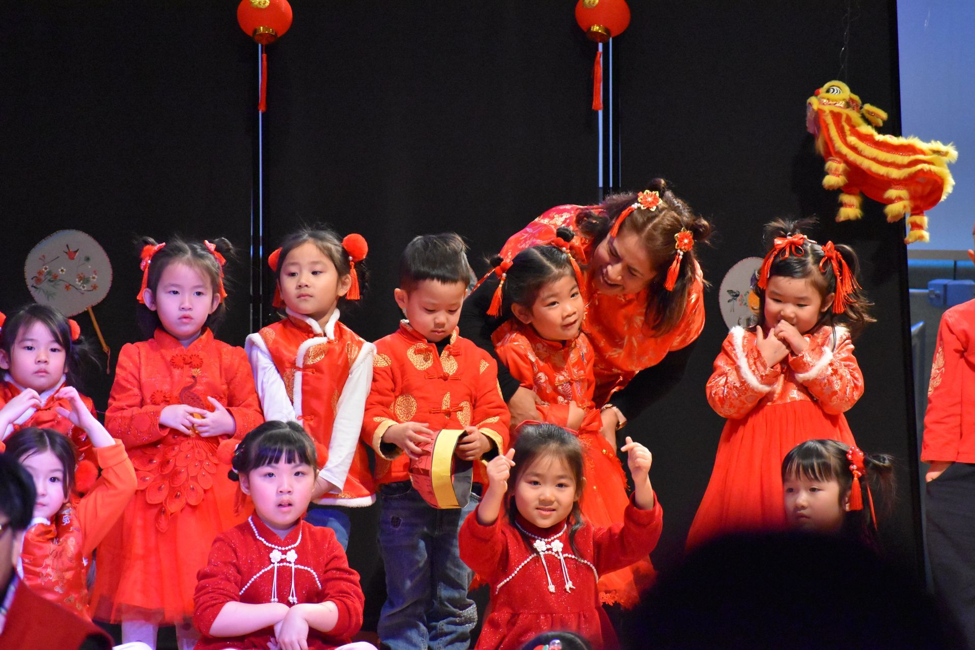 Children in red outfits on stage, some with buns, celebrating; red lanterns and a lion decoration.