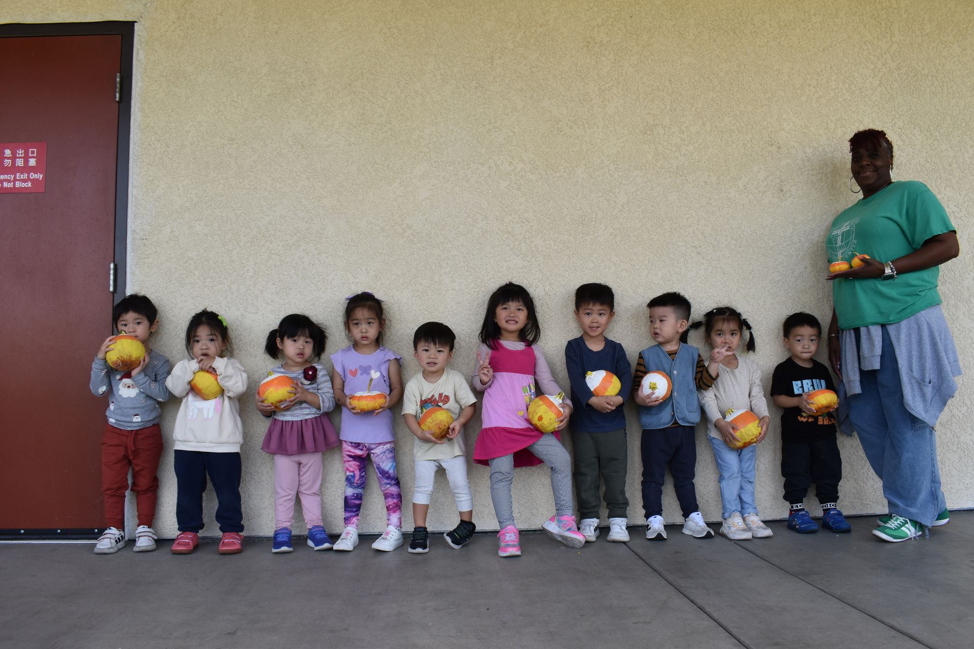 Children and teacher holding balls, standing in front of a tan wall by a door.