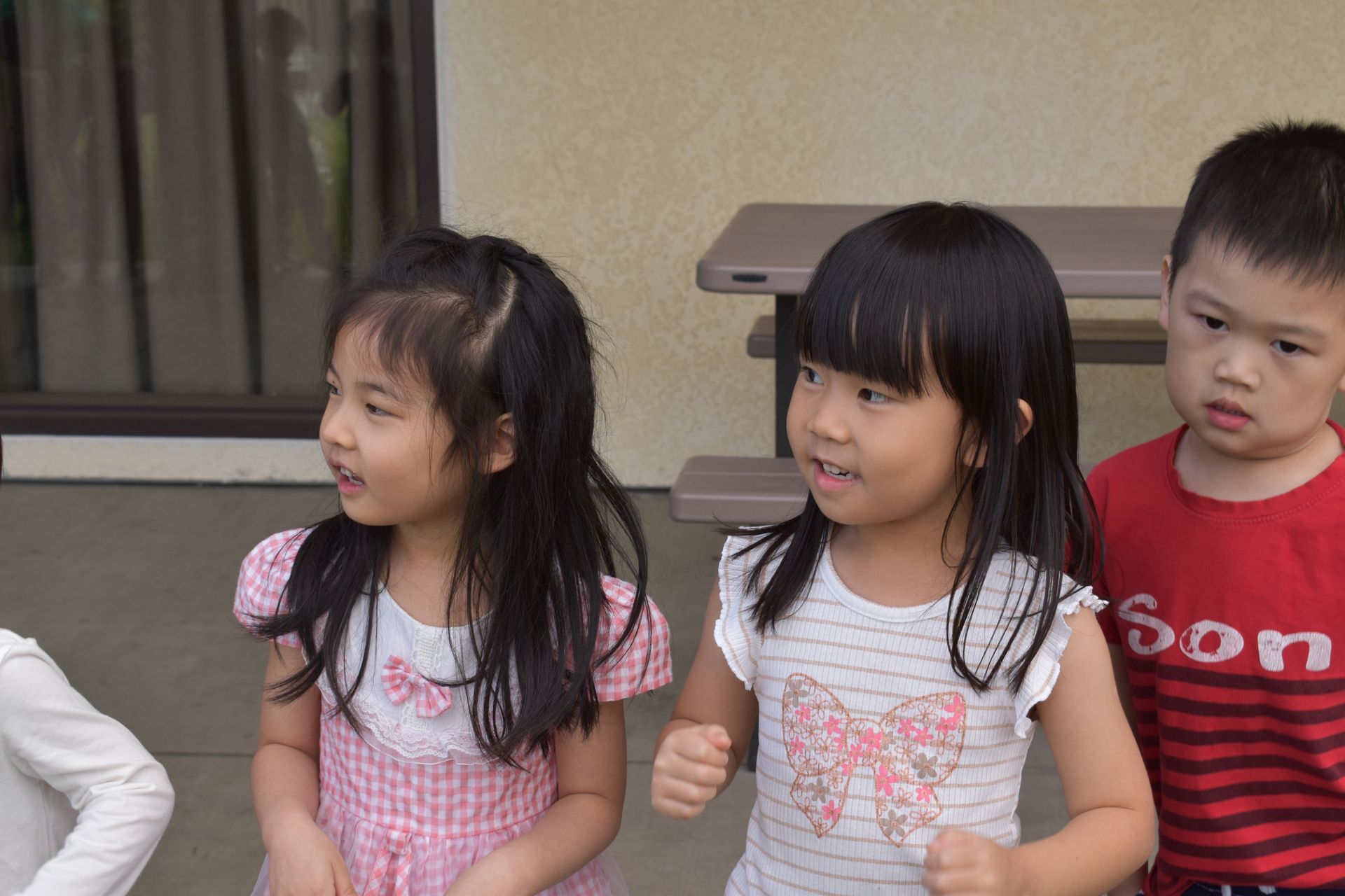 Children standing outside, one wearing a red shirt, looking at something. Two others smiling.