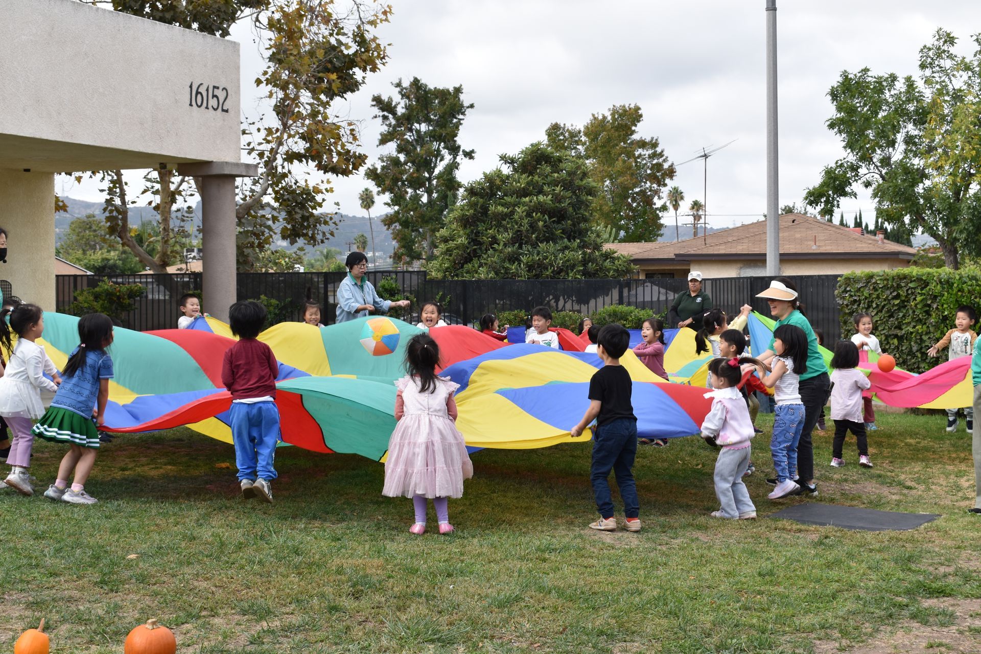 Children playing with a colorful parachute on a grassy lawn; building in background.
