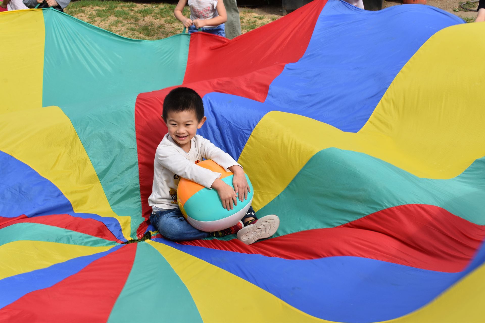 Boy smiling, holding a ball, sitting on a colorful parachute.
