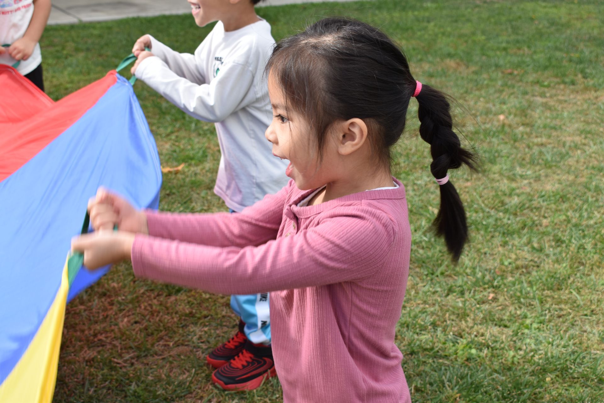 Girl with a braid smiling while holding a colorful parachute in a grassy area with another child.