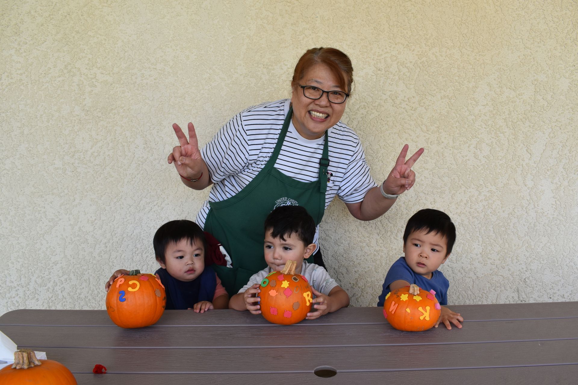 Woman and three children pose behind pumpkins, making peace signs. Table in front.