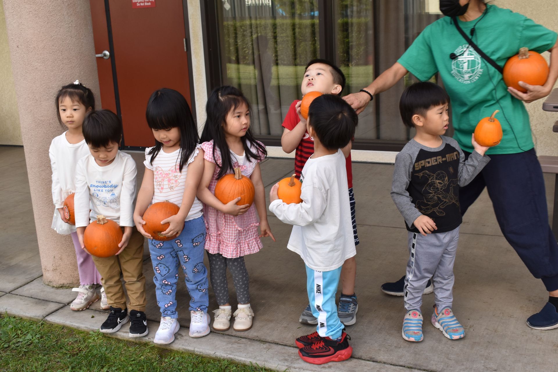 Children holding pumpkins with an adult, outside a building.