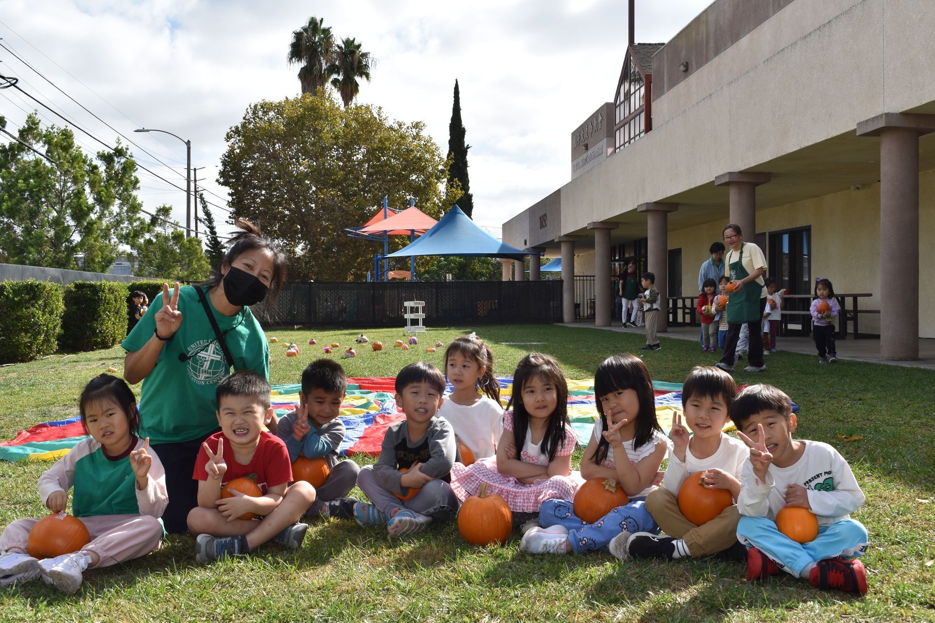 Teacher and children with pumpkins outdoors on a blanket in front of a building.