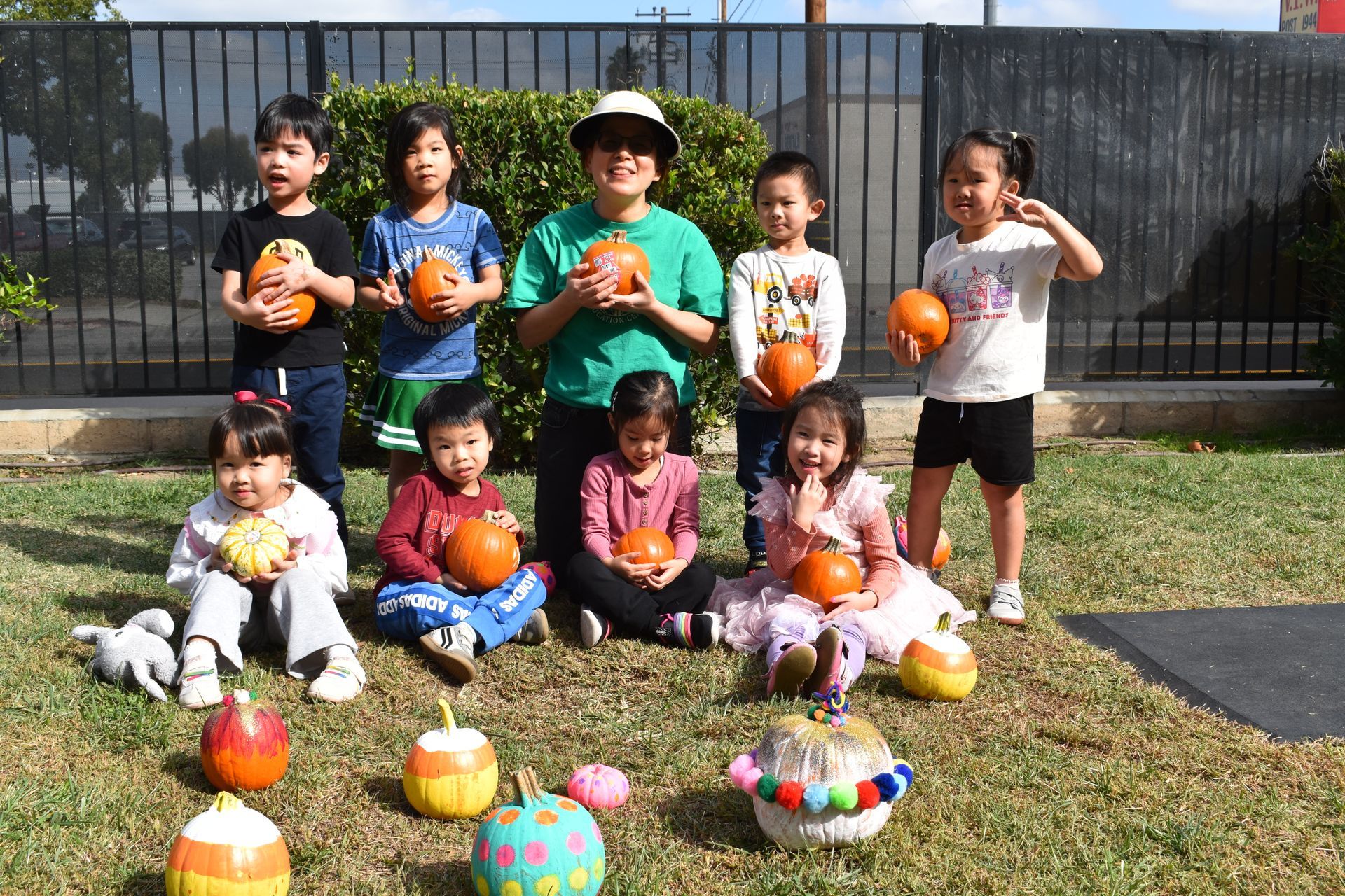 Group of children holding decorated pumpkins in a grassy area near a fence. Some are sitting on the ground, smiling.