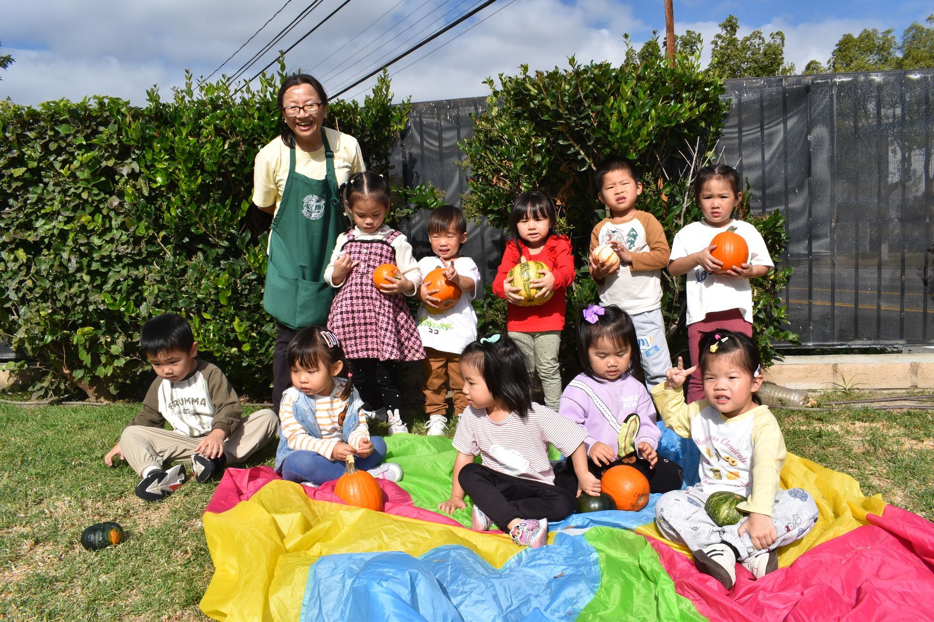 Group of children and teacher with pumpkins on a colorful parachute outside.