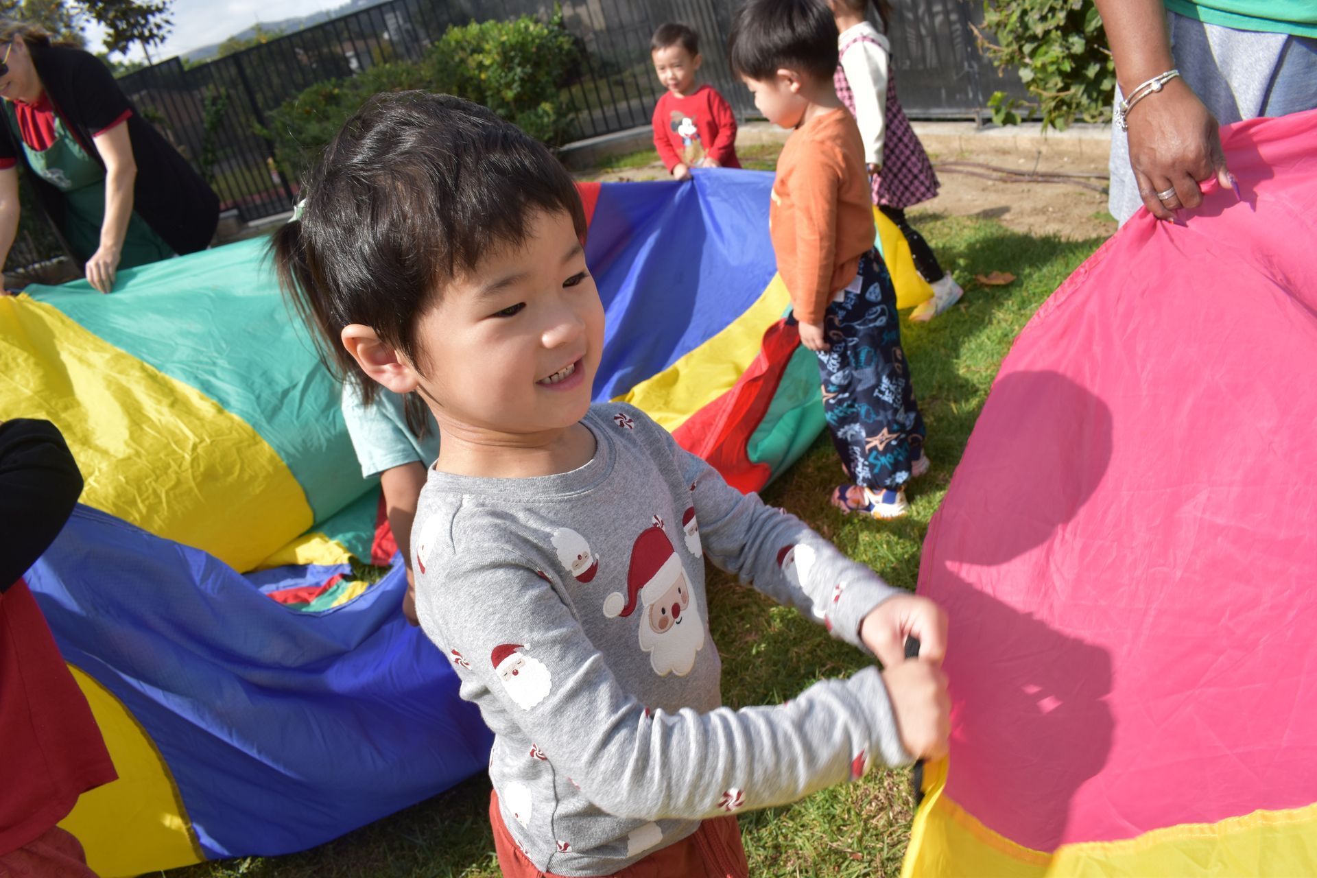 A child smiles, holding a colorful parachute with other kids and adults in an outdoor setting.