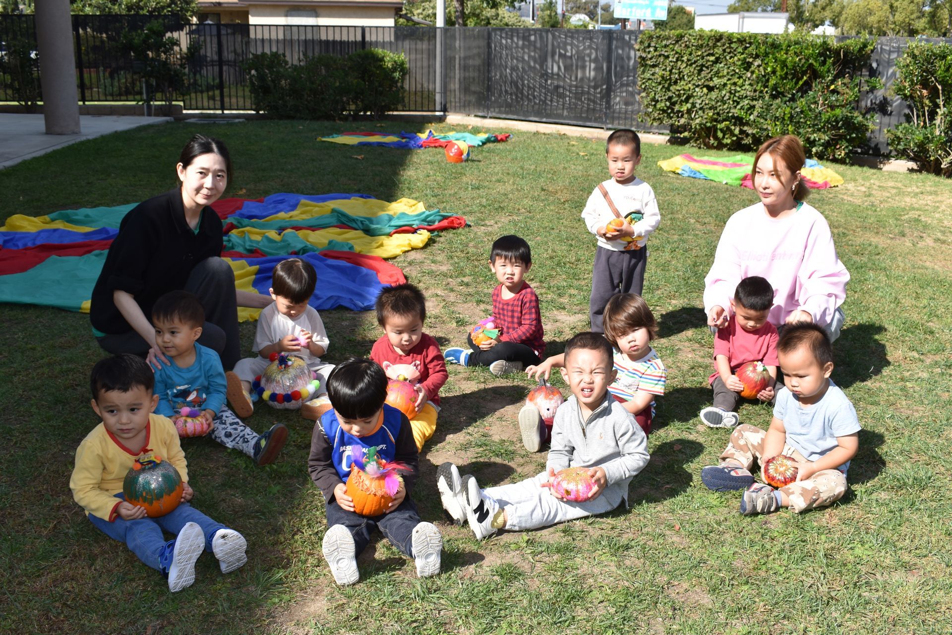 Children and two adults on grass holding pumpkins. Colorful parachute in background. Sunny outdoor setting.