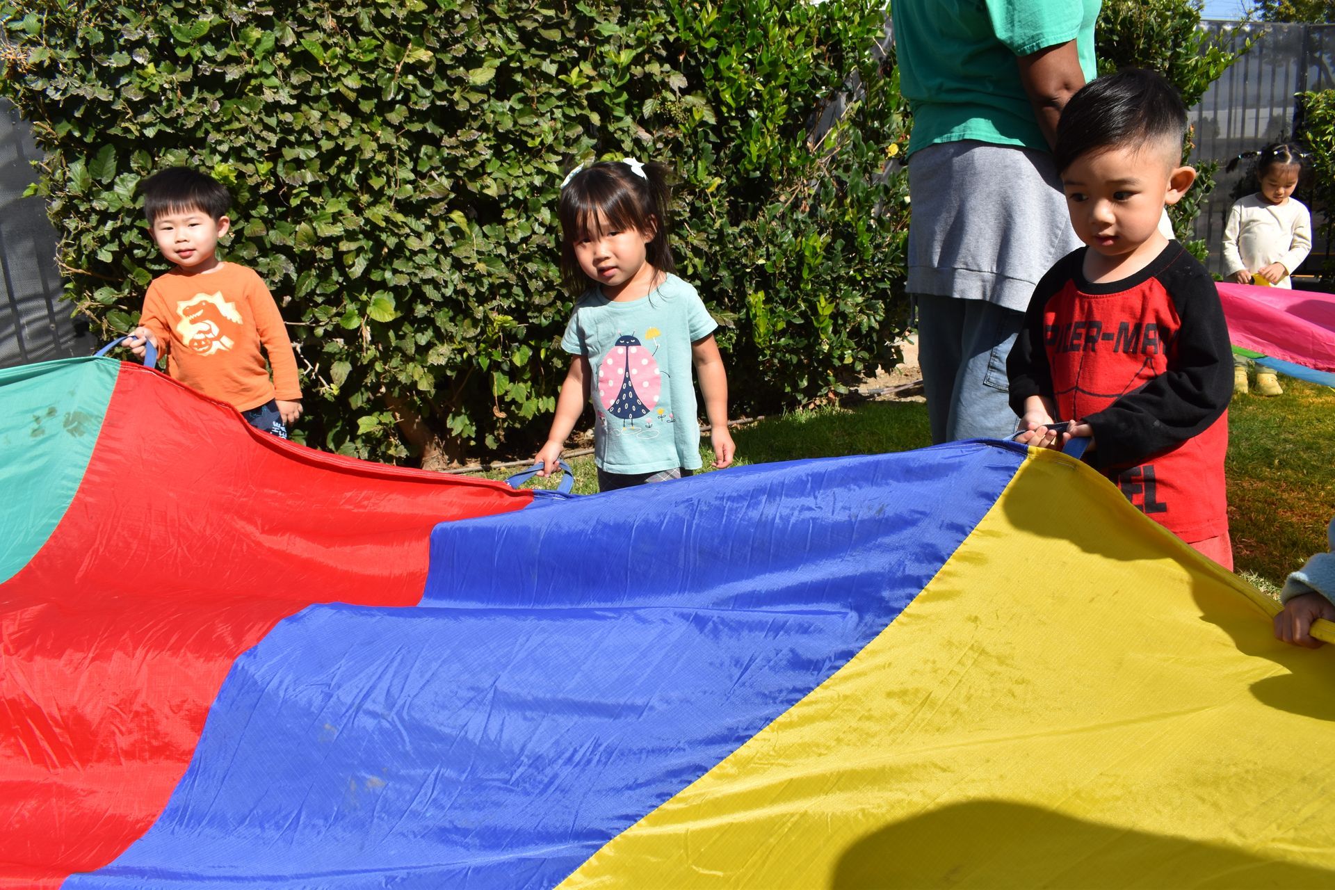 Children holding a colorful parachute outdoors; a person stands behind them.