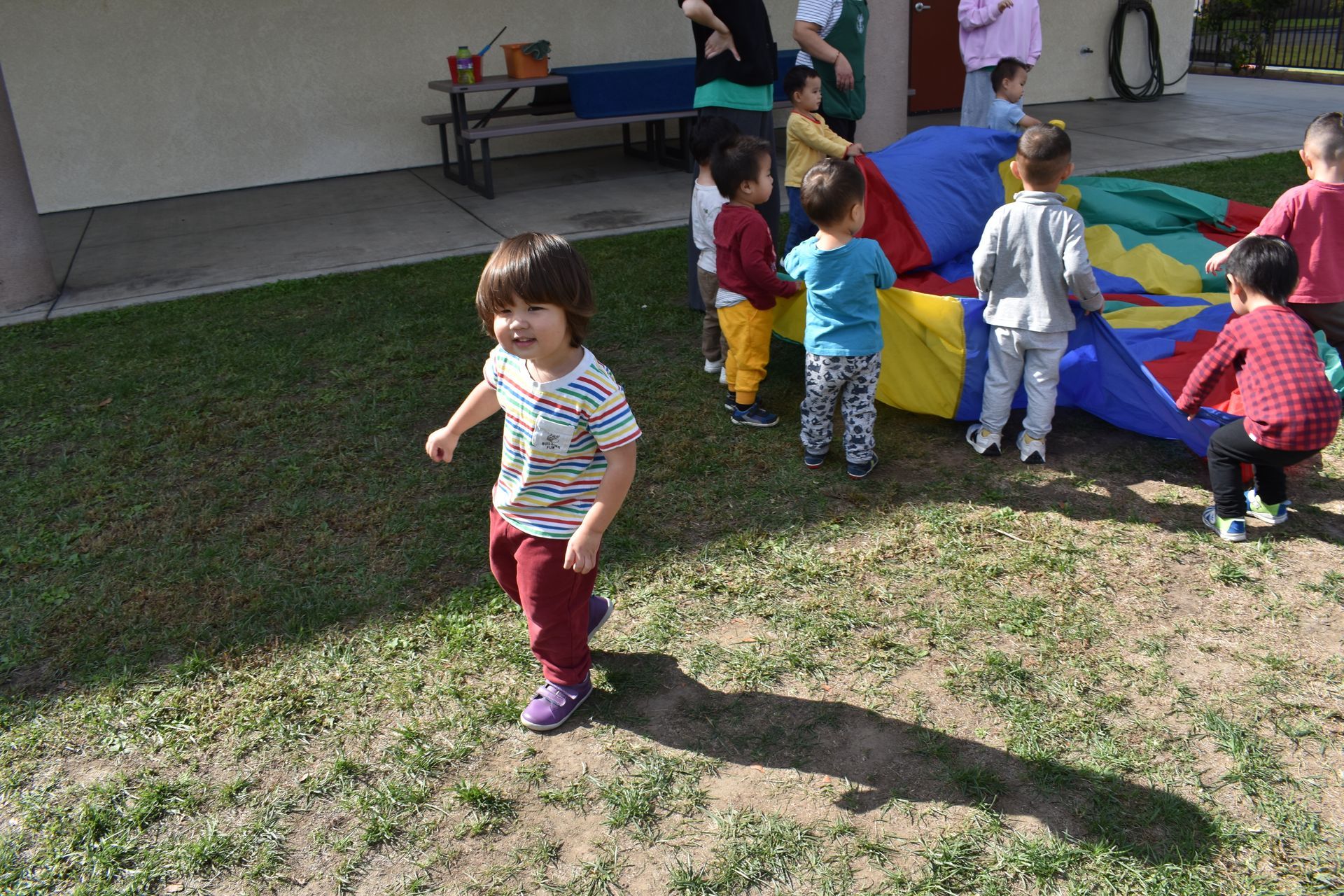 Children playing with a parachute on a grassy area, sunny day. One child running, others holding edges.