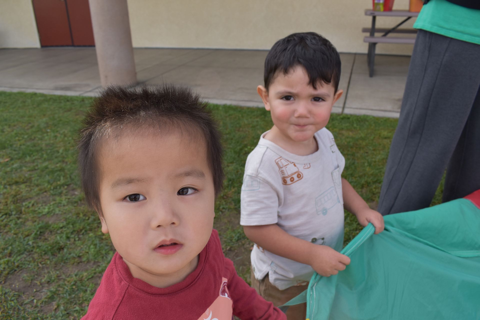 Two children near green fabric outdoors; one looking at the camera, other holding the fabric.