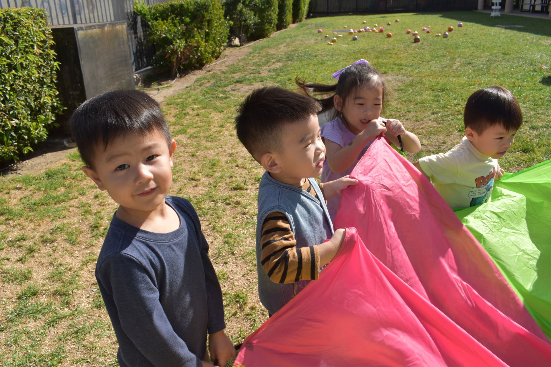 Four children playing with a pink and green parachute on a grassy lawn.