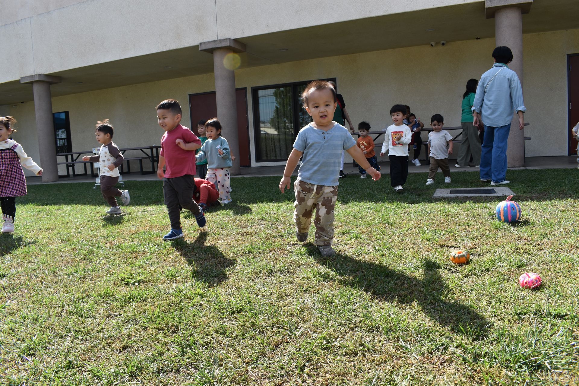 Children playing outside on a grassy area near a building; some run towards the viewer.