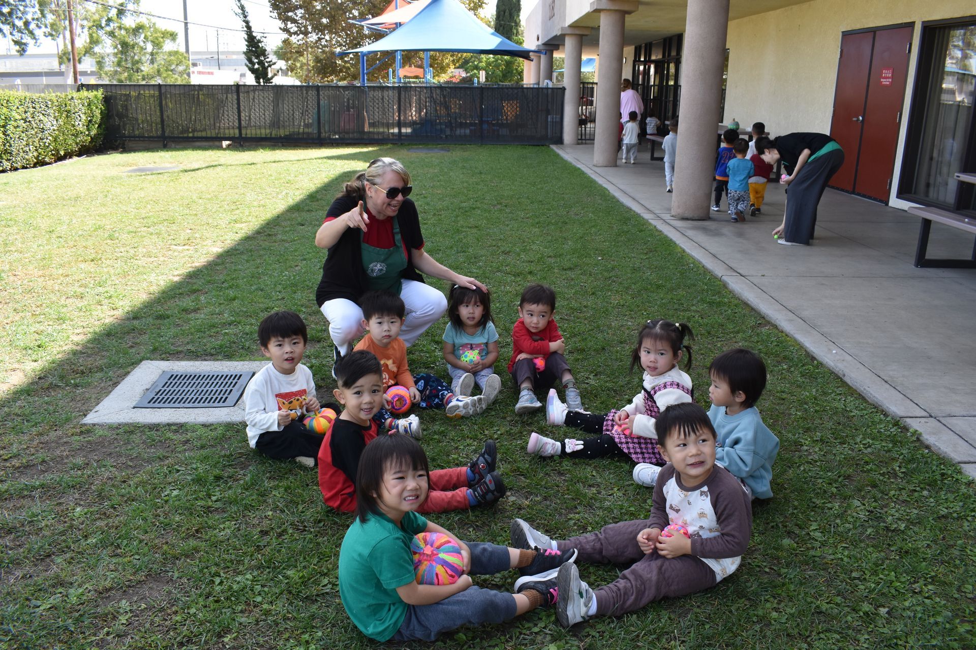 Children sitting in a circle on grass with a teacher outdoors.